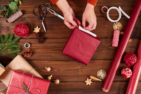 Top view of woman hands decorating red gift box with golden star Packed gifts, spruce branches and boubles on shabby wooden table.