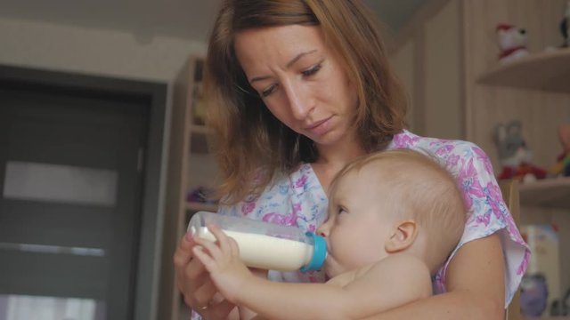 Young mother feeding her baby son with a bottle of milk at home.