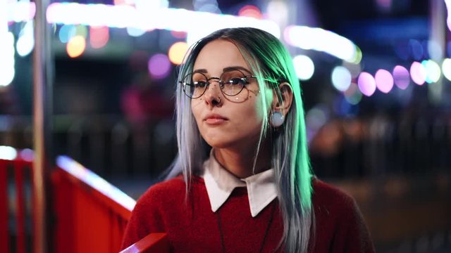 Hipster girl with blue dyed hair and golden sequins as freckles. Woman with nose piercing, transparent glasses, ears tunnels, unusual hairstyle stands in amusement night park