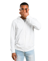 Dark-skinned young man with white sweatshirt covering mouth with hands for saying something inappropriate. Can not speak on isolated white background