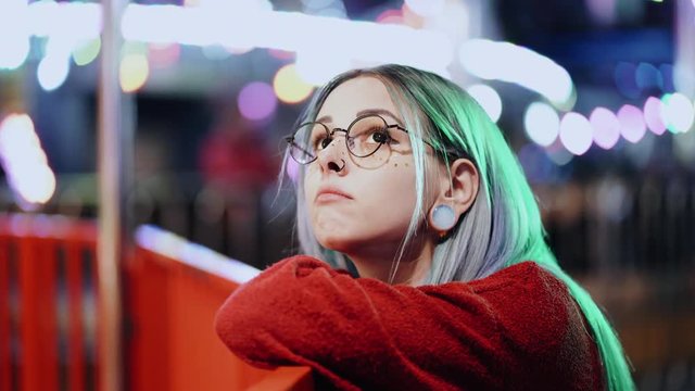 Hipster girl with blue dyed hair and golden sequins as freckles. Woman with nose piercing, transparent glasses, ears tunnels, unusual hairstyle stands in amusement night park