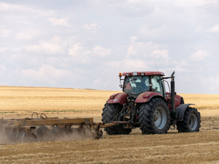 Fototapeta premium Red tractor with plow on harvest field. Summer field and red tractor. Tractor on harvest field. Lowing after harvest. Plowing on the field.