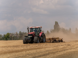 Fototapeta premium Red tractor with plow on harvest field. Summer field and red tractor. Tractor on harvest field. Lowing after harvest. Plowing on the field.