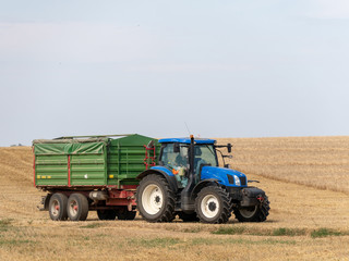 Blue tractor with trailer on the harvest field. Summer field and blue tractor with trailer. Harvest field, blue sky and tractor.
