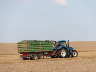 Obraz premium Blue tractor with trailer on the harvest field. Summer field and blue tractor with trailer. Harvest field, blue sky and tractor.
