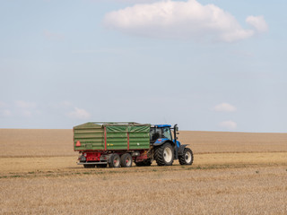 Blue tractor with trailer on the harvest field. Summer field and blue tractor with trailer. Harvest field, blue sky and tractor.