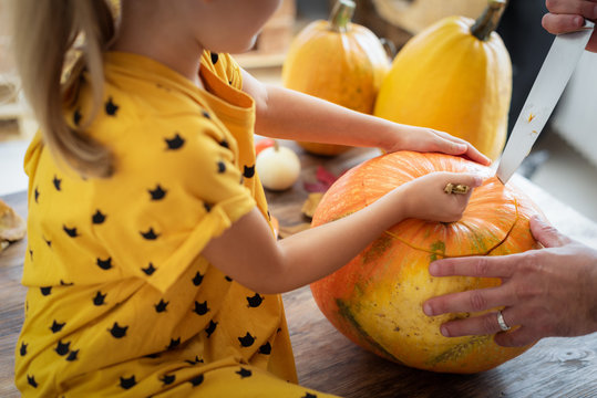 Cute Little Girl Sitting On Kitchen Table, Helping Her Father To Carve Large Pumpkin. Halloween Family Lifestyle Background.