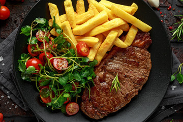 Grilled sirloin steak with potato fries and vegetables, tomato salad in a black plate. rustic table