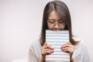 Excited playful Asian girl with dark hair, bites a book , looks nervously at camera, wears...