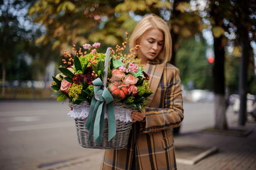 Young blonde woman holding a big wicker basket of flowers against the city