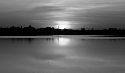 Low light view with black and white of quiet freshwater lake is reflection of the sun on water surface. Silhouetted the trees along the coast. During the sunset.