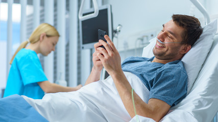 Recovering Patient Uses Smartphone while Lying on a Bed in the Hospital. Friendly Nurse Comes in to Check on Him.