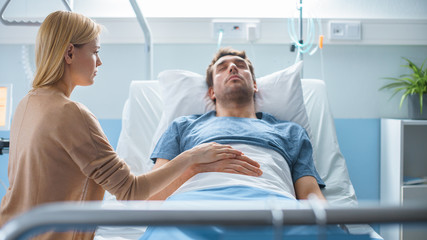 Obraz premium In the Hospital Sick Man Lying on the Bed, His Visiting Wife Hopefully Sits Beside Him Holds His Hands and Hopes for Recovery.