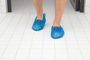 Man's shoes in blue overshoes on the white tiles floor. Patient or visitor going at hospital. Front view. Close up.