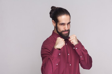 Portrait of serious handsome man with dark collected long hair and beard in red shirt standing, looking at camera with boxing fists and ready to attack. indoor studio shot, isolated on gray background