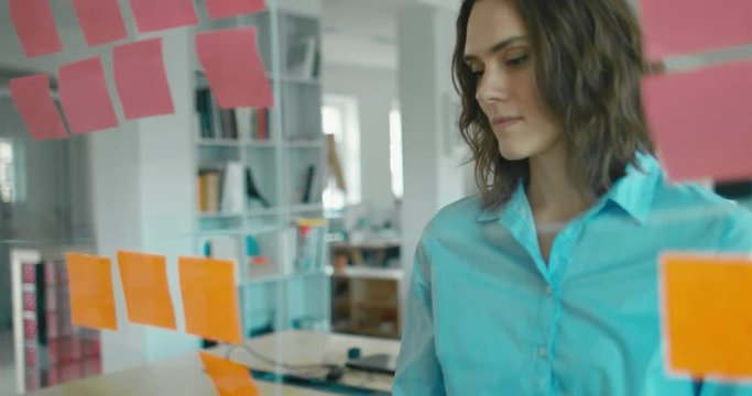 Caucasian colleagues standing near glass wall with sticky notes, framework for managing work, scrum methodology. 4K UHD 60 FPS SLOW MOTION