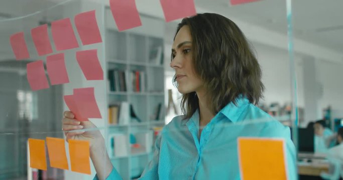 Caucasian female worker standing near glass wall with sticky notes, framework for managing work, scrum methodology. 4K UHD 60 FPS SLOW MOTION