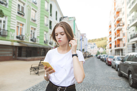 Portrait of a serious stylish girl standing on the street in the town, looking at the smartphone in her hands and listening to music in the headphones.