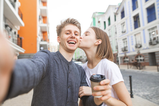 Portrait of a lovers of a young couple takes selfie on the street of the town. Smiling young man with a cup of coffee in his hands takes selfie, his girlfriend kisses on his cheek. Love story
