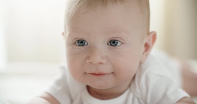 A cute happy baby lying on white bed sheet, looking around and happily laughing and smiling. closeup 4k