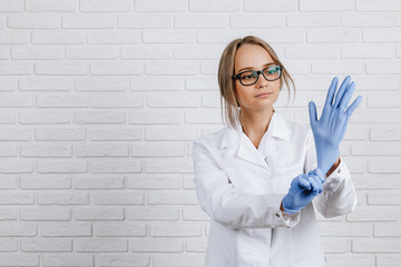 Doctor woman wearing black glasses in a white coat gently puts on her hands blue gloves on a white background