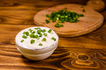 Glass bowl with sour cream and cutting board with chopped green onion on wooden table