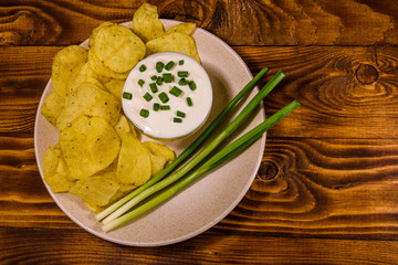 Ceramic plate with potato chips and glass bowl with sour cream on wooden table. Top view