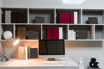 Homemade workplace, a computer is standing on a table, a desk lamp is shining on the background of a shelf with books and photo frames