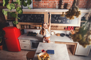Attractive blonde long hair girl wearing sleapwear dancing below the music in the red loft comfortable sunny kitchen, raising hands and happy of the new day