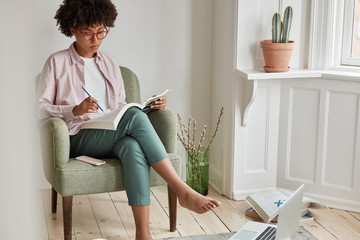 Shot of beautiful black freelancer with Afro hairstyle, sits in comfortable armchair, reads...