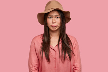 Indoor shot of dissatisfied woman has placid discontent facial expression, purses lips in displeasure, being in low spirit, dressed in fashionable hat, poses against pink background. Negative feeling
