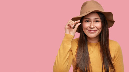 Positive girl shows something invisible and very little, gestures with hand, smiles gladfully, wears stylish hat, has freckled skin, poses against pink background with free space for your promotion
