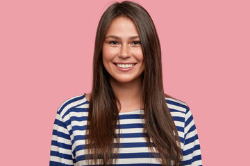 Brunette satisfied girl with pleasant toothy smile, shows natural beauty, wears casual striped sailor sweater, being in good mood, has casual talk with close person, stands against pink background - obrazy, fototapety, plakaty