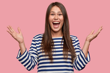 Waist up shot of happy girl with freckled skin, raises hands, gestures positively, screams with joy, glad to complete task and overcome hardships dressed in oversized striped sweater, isolated on pink