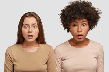 Horizontal shot of diverse women gaze at camera with surprised expressions, stand closely to each other, dressed in casual clothes, recieve unexpected news from groupmate or colleague, stand indoor
