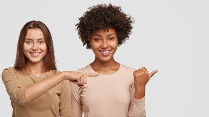 Photo of satisfied interracial young women point aside with joyful expressions, show nice place to...