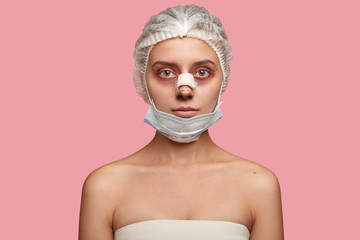 Shot of serious woman wears medical hat and mask, has rhinoplasty, preapres for blepharoplasty, has...