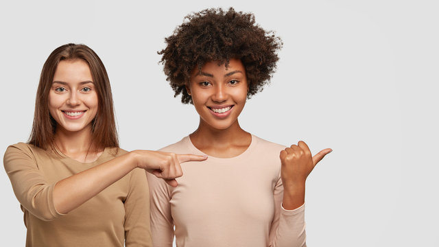 Photo of satisfied interracial young women point aside with joyful expressions, show nice place to visit, have tender smiles on faces, stand closely to each other, isolated over white background.