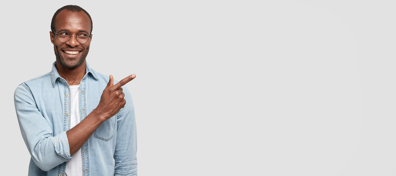 Medium length shot of attractive black man smiles brightfully, dressed in fashionable denim shirt, points at white wall with index finger, advertises something not expensive and of good quality