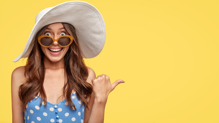 Studio shot of pleasant looking gorgeous young lady in trendy white hat, shows you blank space or direction to resort place, has toothy smile, poses over yellow background. Advertisement concept - obrazy, fototapety, plakaty