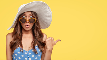 Stunned beautiful young European woman with dark wavy hair, scared expression, dressed in fashionable outfit, ready for summer vacation, indicates aside at blank copy space, yellow background - obrazy, fototapety, plakaty