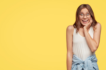 Photo of satisfied young Caucasian woman smiles positively, keeps hand on chin, being in good mood, notices desirable thing, poses against yellow background with free space for your promotion