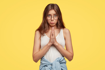 Obraz premium Candid shot of miserable guilty girlfriend asks for forgiveness, purses lips, keeps palms pressed together over chest, wears casual t shirt, denim jacket around waist, isolated over yellow wall