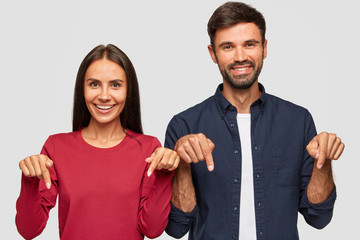 Horizontal shot of cheerful Caucasian young adult woman and man point with index fingers down,...