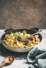 Meat dinner. Braised beef meat with potato and carrot with fresh parsley in cooking pan over white marble table background, selective focus, copy space. Comfort winter food