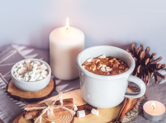 A cup of hot cocoa with marshmallows and candles on a table surrounded by winter or Christmas decor, closeup