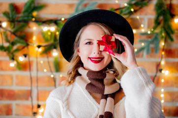 Portrait of a young cozy woman in white sweater with present gift and Christmas lights and pine branch