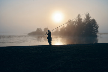 Silhouette of fisherman fishing with spinning rod on river bank at misty foggy sunrise. Isolated outdoor portrait of man standing by lake at sunset holding fishing rod and camp chair under his arm