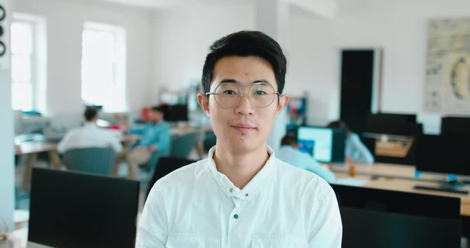 Portrait of young adult mid Asian Chinese male smiling and looking into camera, busy office in the background. 4K UHD 60 FPS SLOW MOTION