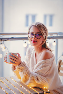 Young woman with cup of coffee sitting at table in near fairy lights in evening time.
